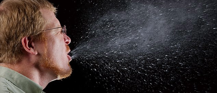 Man sneezing in front of a black background with visible droplets illustrating how infections can spread through the air.